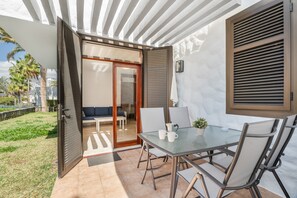 Une terrasse ouverte avec une table en verre, des chaises et une porte-fenêtre coulissante donnant sur un espace de détente intérieur.
