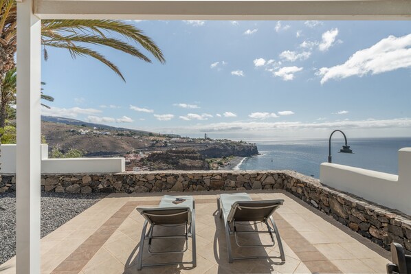 A terrace with two chairs and a table, overlooking a coastal landscape with a stone wall and a view of the sea.