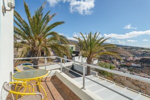A balcony with a glass table and chairs, palm trees, and a view of a town.
