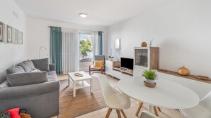 En moderne stue med sofa, spisebord, stole, et tekøkken med tv og et vindue med gardiner.