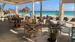 An outdoor seating area with tables and chairs, a thatched roof, and a view of the beach and ocean. - Private Beach | Terrace and Pool • Fast WiFi (Punta Cana)