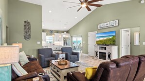 A spacious living room with a brown leather sofa, a wooden coffee table, and a television. A ceiling fan and a sign reading "SAND AND SURF" are visible. - 🏖️Nirvana in Navarre | Stunning 3BR Home w/ Private Pool + Walk to Beach🏖️ (Navarre)