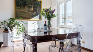 A dining area with a glass table, clear chairs, a vase with flowers, a wine bottle, and a painting on the wall. - Saint Peter's Luxury Terrace Suites (Roma)