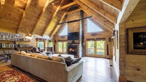 A spacious loft with a high ceiling, exposed wooden beams, a stone fireplace, a large sofa, and a bookshelf filled with books.