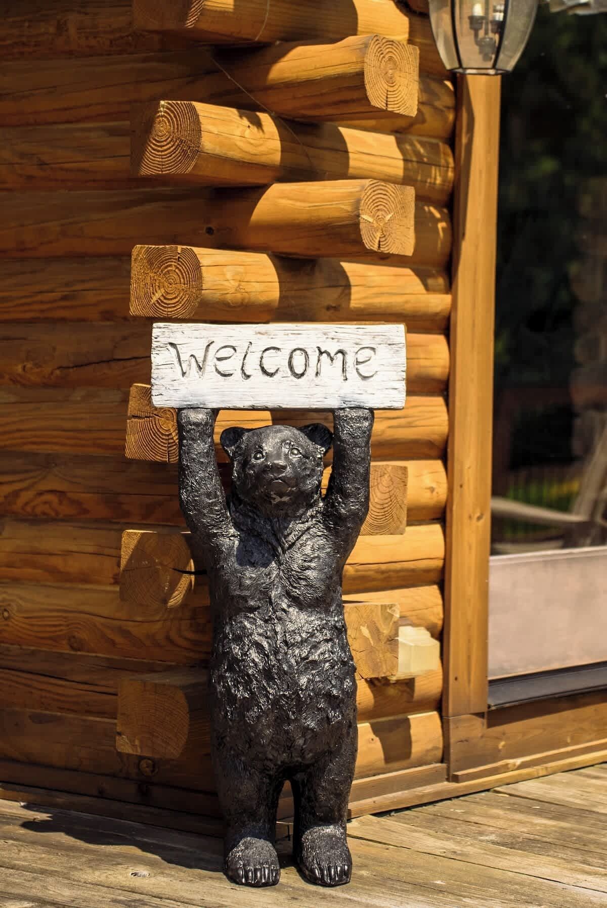 A bear holding a 'Welcome' sign in front of a wooden log cabin.