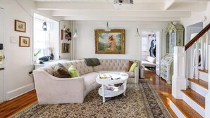 Una sala de estar con un sofá blanco, una mesa de centro y un tapete estampado. Se ve una escalera a la derecha y una puerta al fondo.