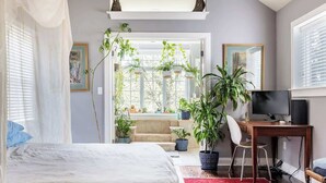 A bedroom with a bed, a desk, a chair, and a TV. There are plants and a window. - Unique Luxury Central Historic Landmark Home (Lambertville)
