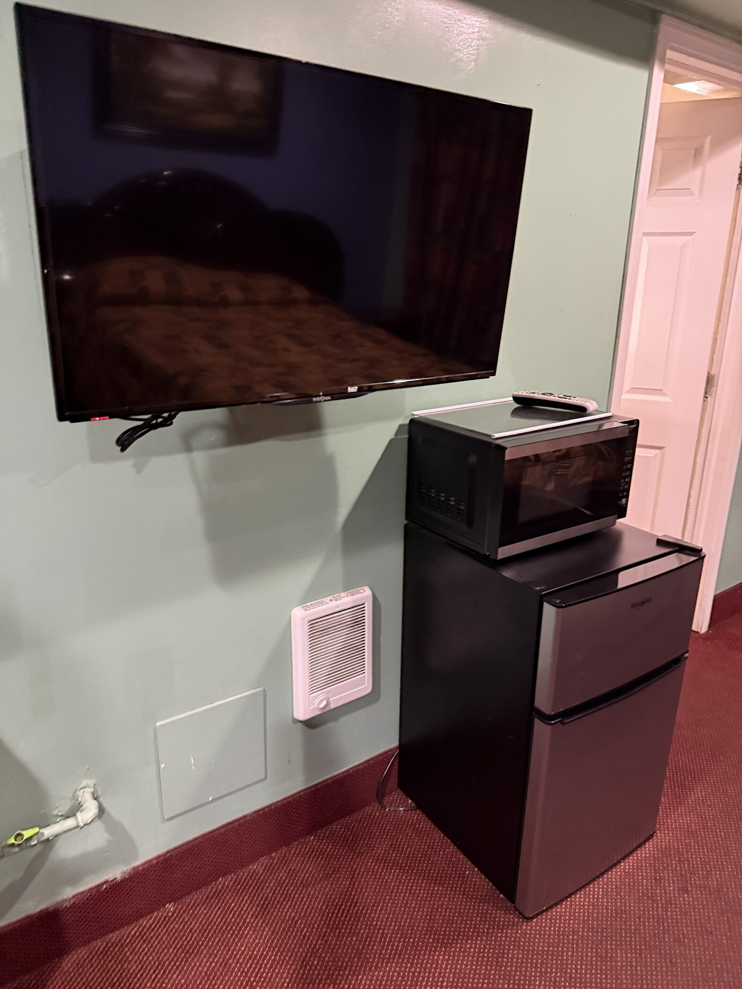 A room with a flat-screen TV mounted on the wall, a black refrigerator, and a microwave on top of it.