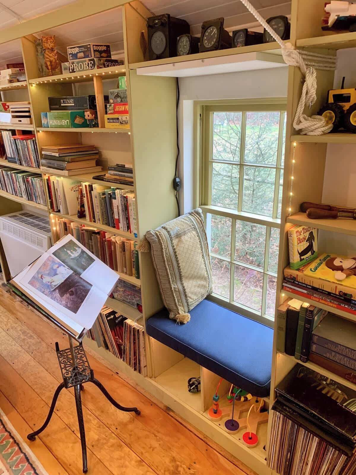 A cozy reading nook with a bench and a window, surrounded by bookshelves filled with various books and puzzles.