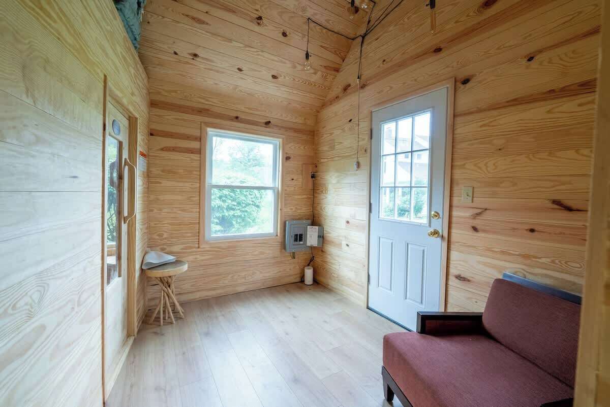 A wooden cabin interior with two windows, a door, a small table, and a bench.