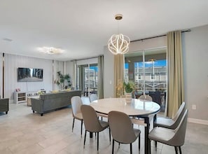 Ein modernes Wohnzimmer mit einem runden Esstisch, grauen Sofas und einem großen Fenster mit Vorhängen.