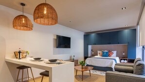 Een moderne hotelkamer met een kitchenette, een bed met blauw hoofdeinde, en een zithoek met een bank en fauteuils.