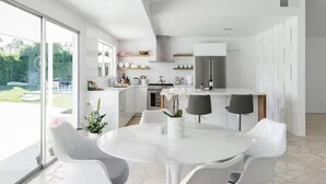 A modern kitchen with white cabinetry, a marble island, and a dining area with white chairs and a table. - Besveca House - Modern Zen (Palm Springs)