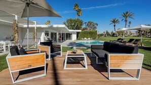 A modern outdoor seating area with white and wooden furniture, a pool, and palm trees in the background. - Besveca House - Modern Zen (Palm Springs)