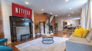 Una sala de estar con chimenea, una escalera, un sofá con cojines amarillos y un cartel que dice 'NETFLIX'.