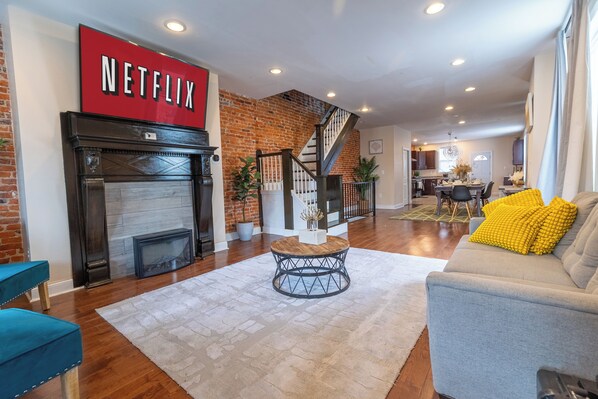 A living room with a fireplace, a staircase, a sofa with yellow cushions, and a sign that says 'NETFLIX'.