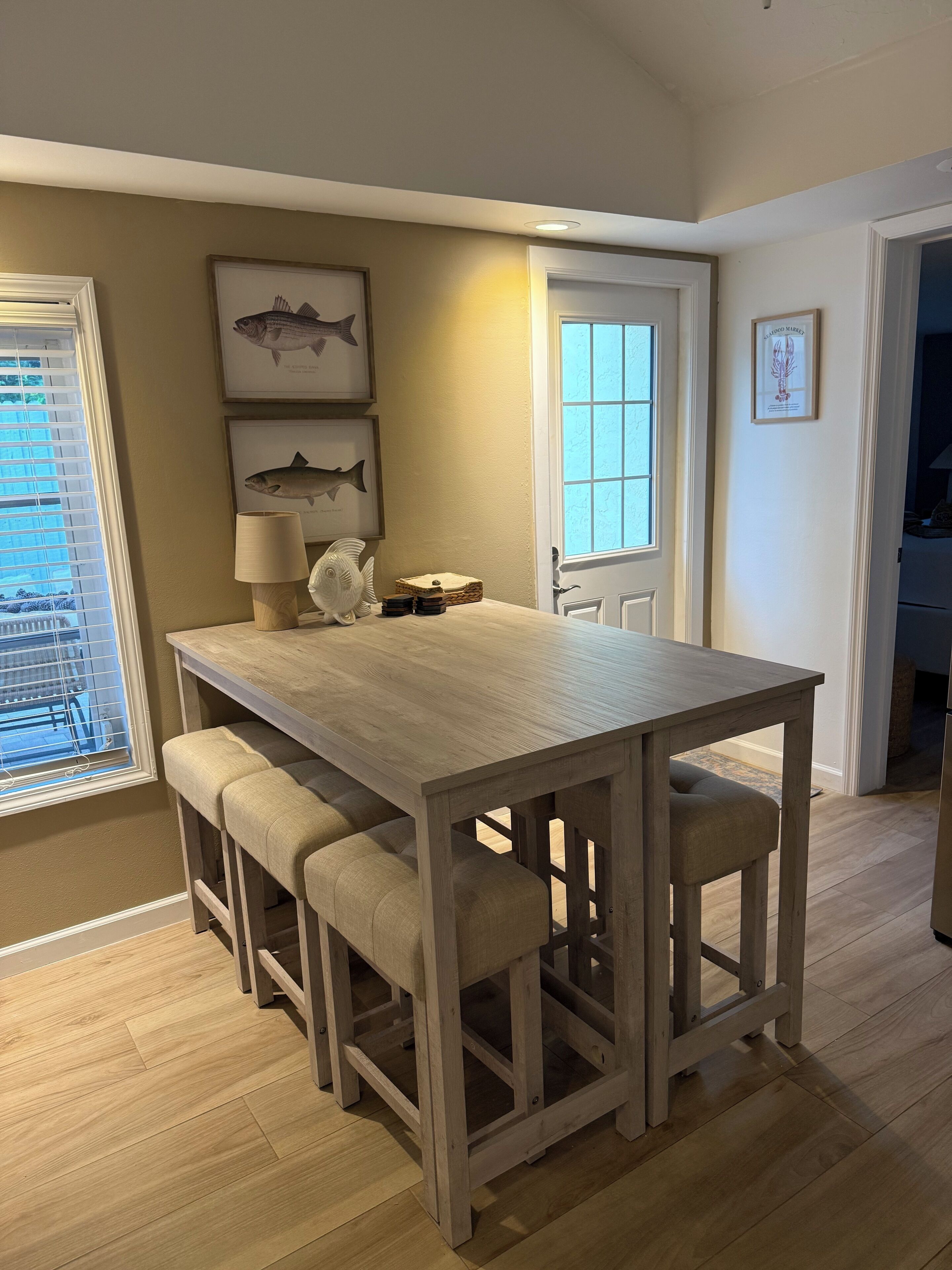 A dining area with a wooden table and bench seating, framed fish artwork on the wall, and a window with blinds.