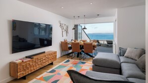 A modern living room with a large TV, a wooden console table, a gray sofa, and a dining area with a view of the sea. - 1 Moonrakers - Three Bedroom House, Sleeps 6 (Carbis Bay)