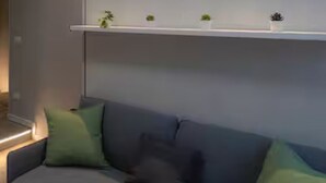 Ett modernt vardagsrum med en grå soffa, gröna och svarta kuddar och en vit hylla med små växter.