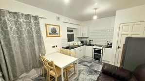 A compact kitchen with white cabinets, a dining table with chairs, a refrigerator, and a black leather sofa. - Modern 2 Bed Flat - Ideal for Families  (Ashton-under-Lyne)