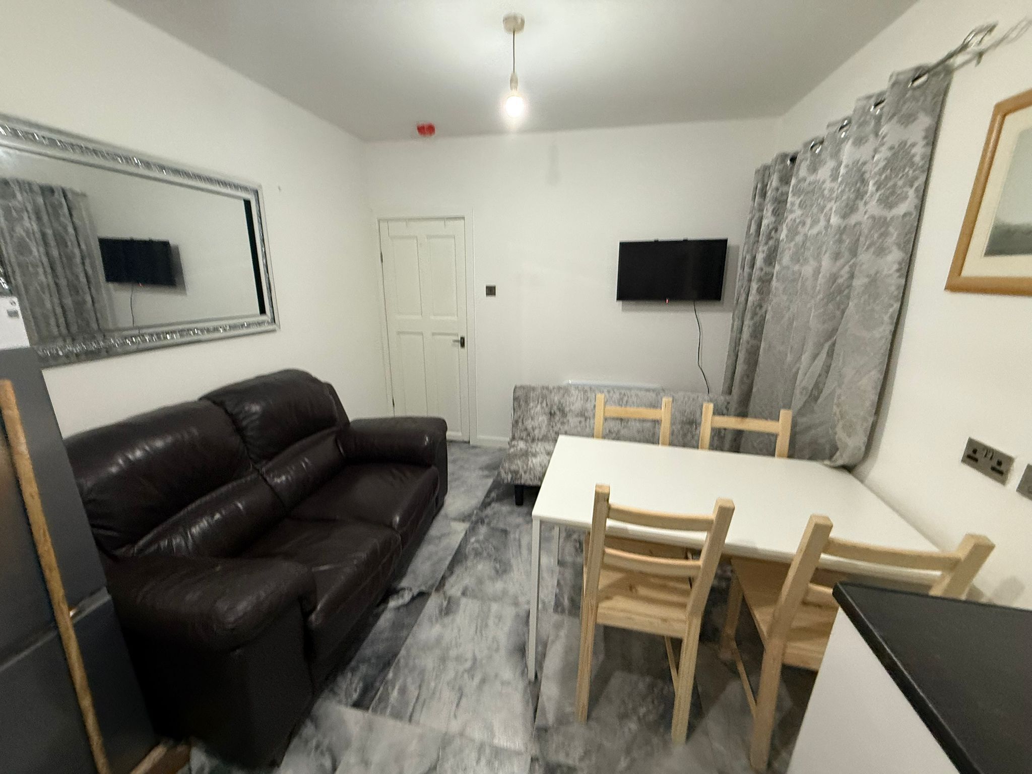 A small, compact living space with a sofa, a dining table with chairs, a flat-screen TV mounted on the wall, and a door.