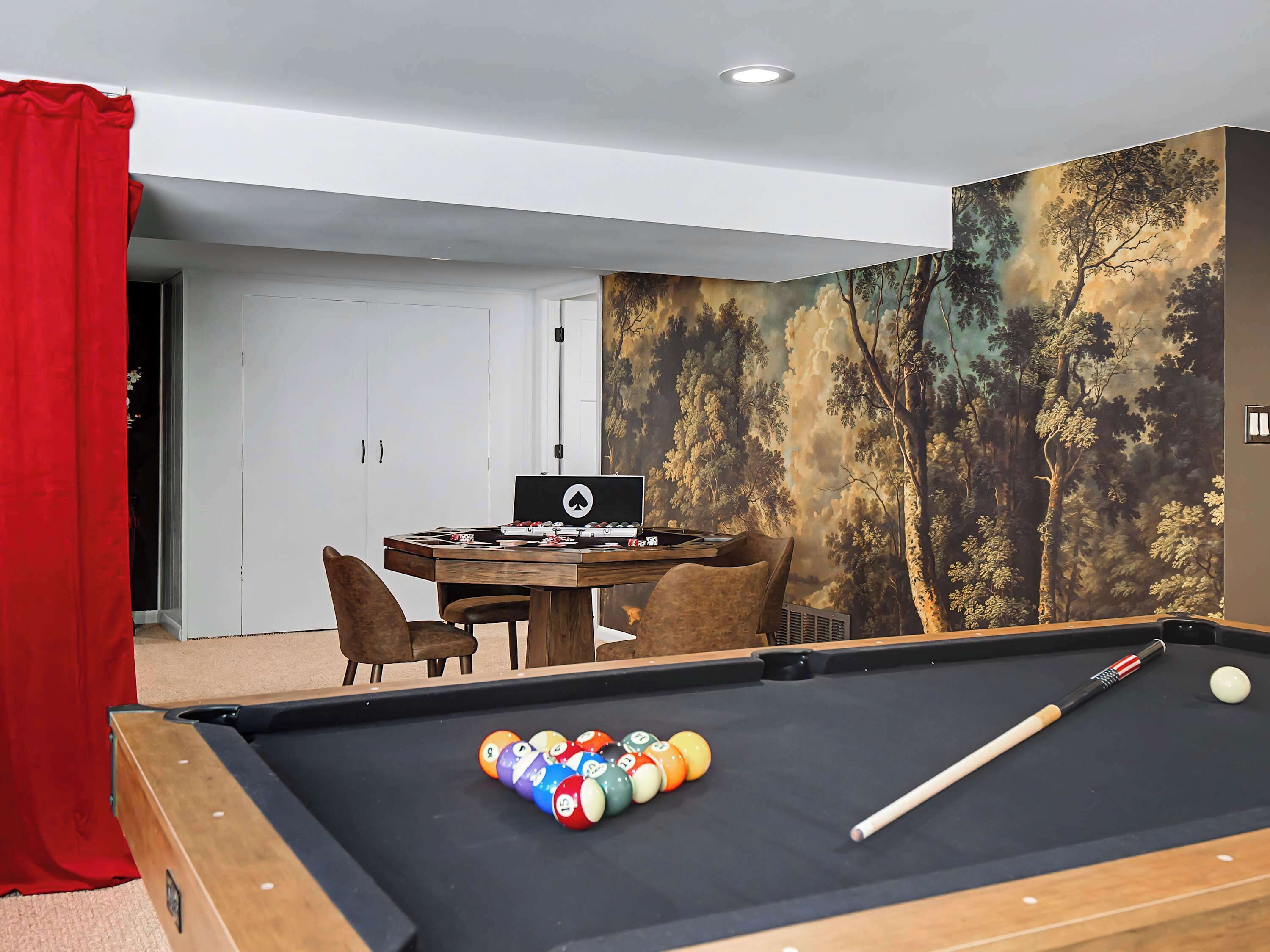 A pool table with a cue stick and billiard balls, a dining table with chairs, and a large wall mural of a forest scene.