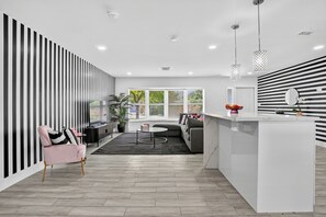 Ein modernes Wohnzimmer mit einer Couch, einem rosa Sessel, einem Couchtisch und einer Küchenzeile mit weißer Kochinsel und einer Pendelleuchte.