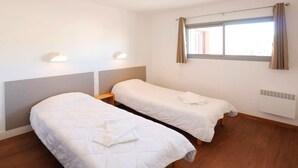 Une chambre avec deux lits simples, une fenêtre avec des rideaux, un radiateur fixé au mur et une petite table avec une lampe.