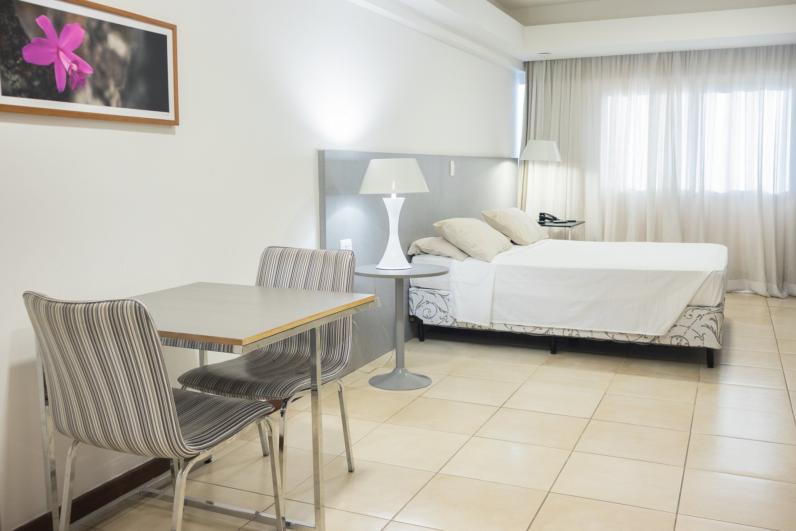 A hotel room with a bed, a desk, a chair, a lamp, and a framed picture on the wall.