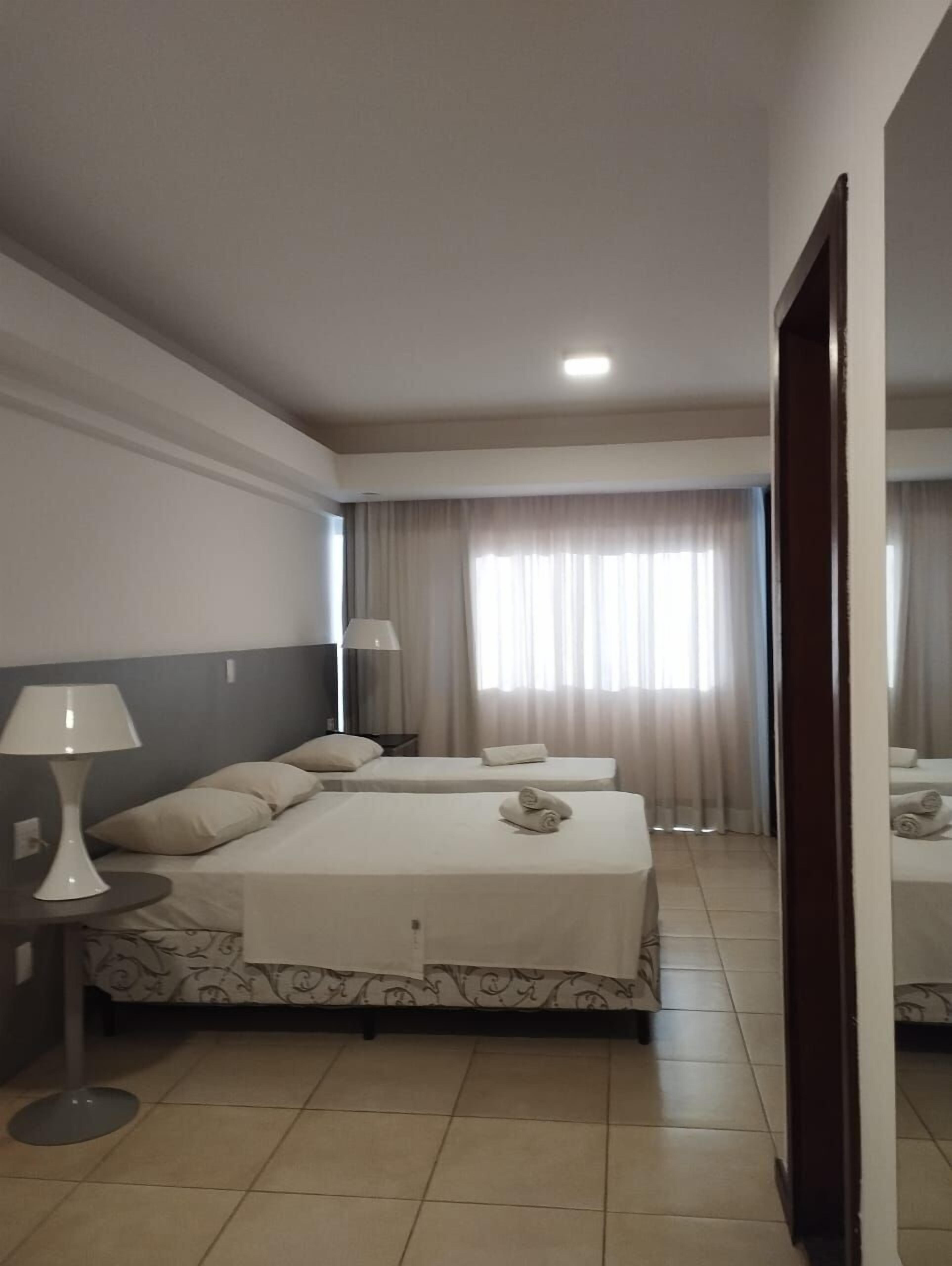 Habitación de hotel con cama, dos mesitas de noche, lámpara y ventana con cortinas.
