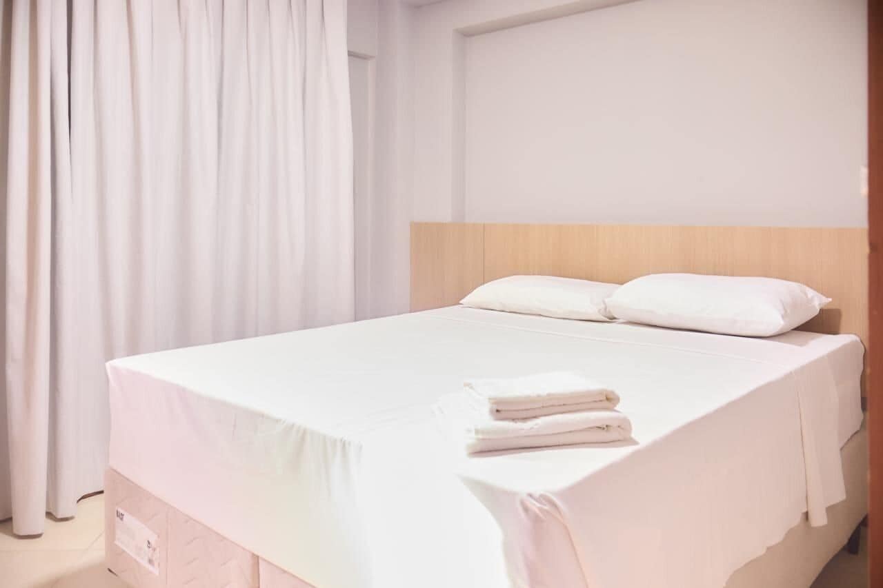 Un lit bien fait, avec du linge de lit blanc et des oreillers, une tête de lit en bois et un rideau sur le côté gauche.