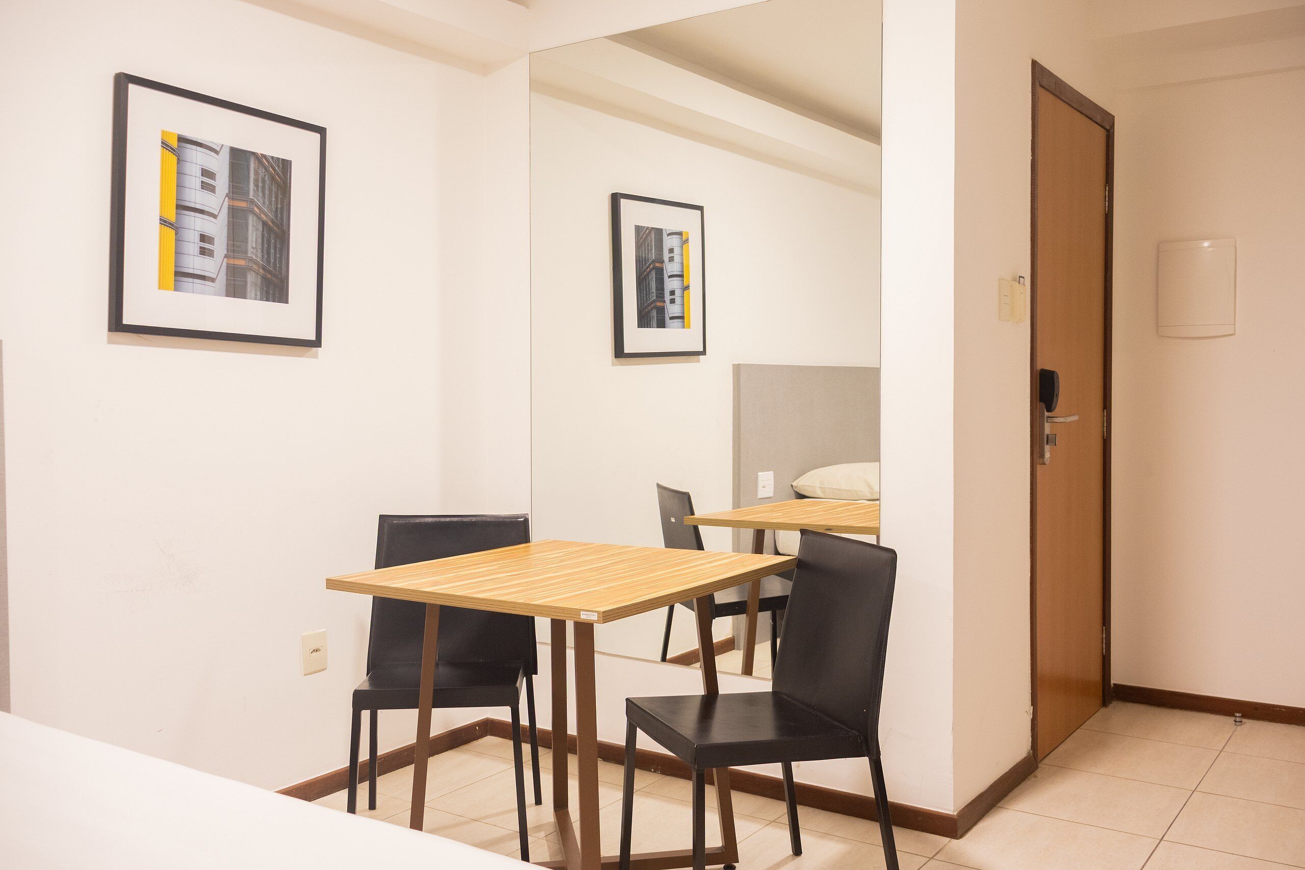 A hotel room with a wooden table and two black chairs, a door, and two framed pictures on the wall.