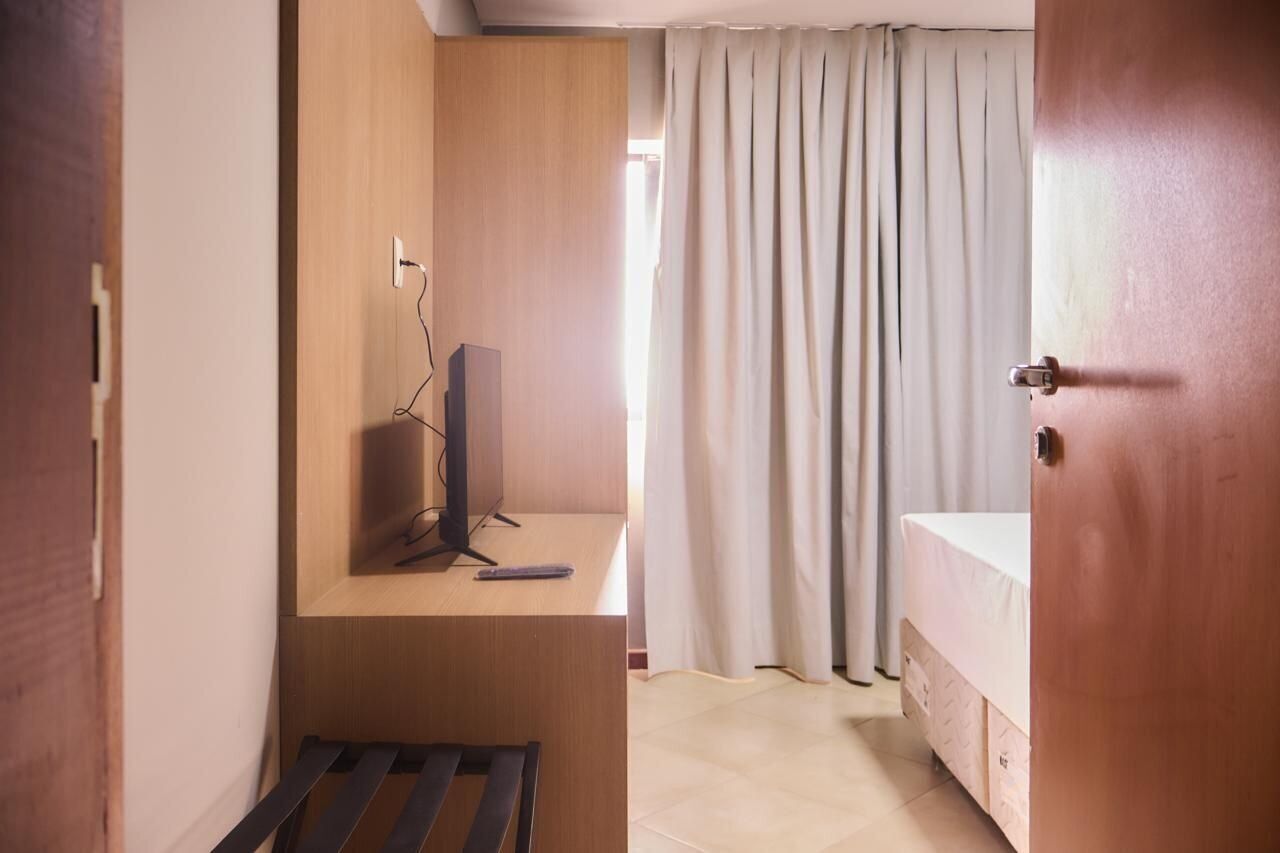 A compact room with a wooden desk, a television, a bed, and a window with curtains.