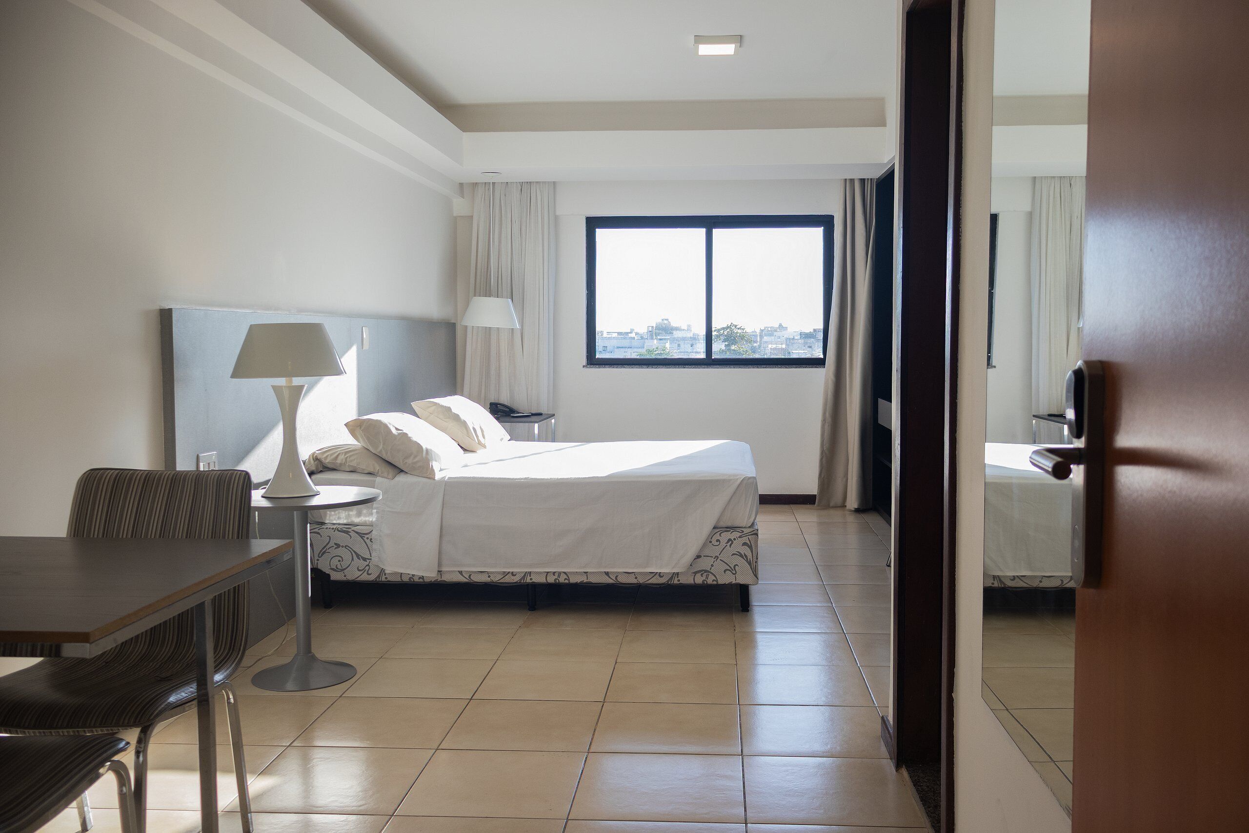 A hotel room with a bed, a desk, a chair, a lamp, and a window with a city view.