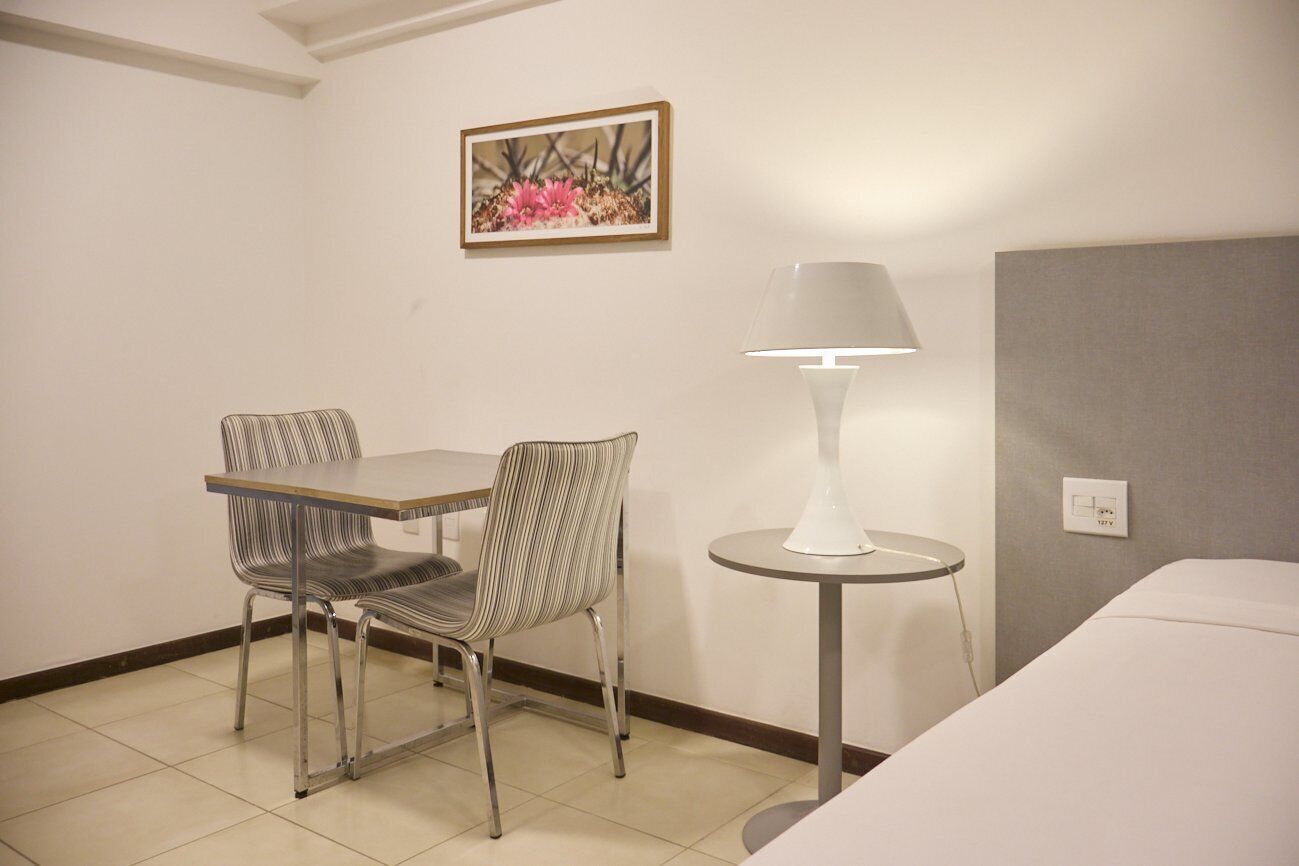 Una habitación de hotel con una cama, una mesita redonda con una lámpara, dos sillas a rayas y un cuadro en la pared.
