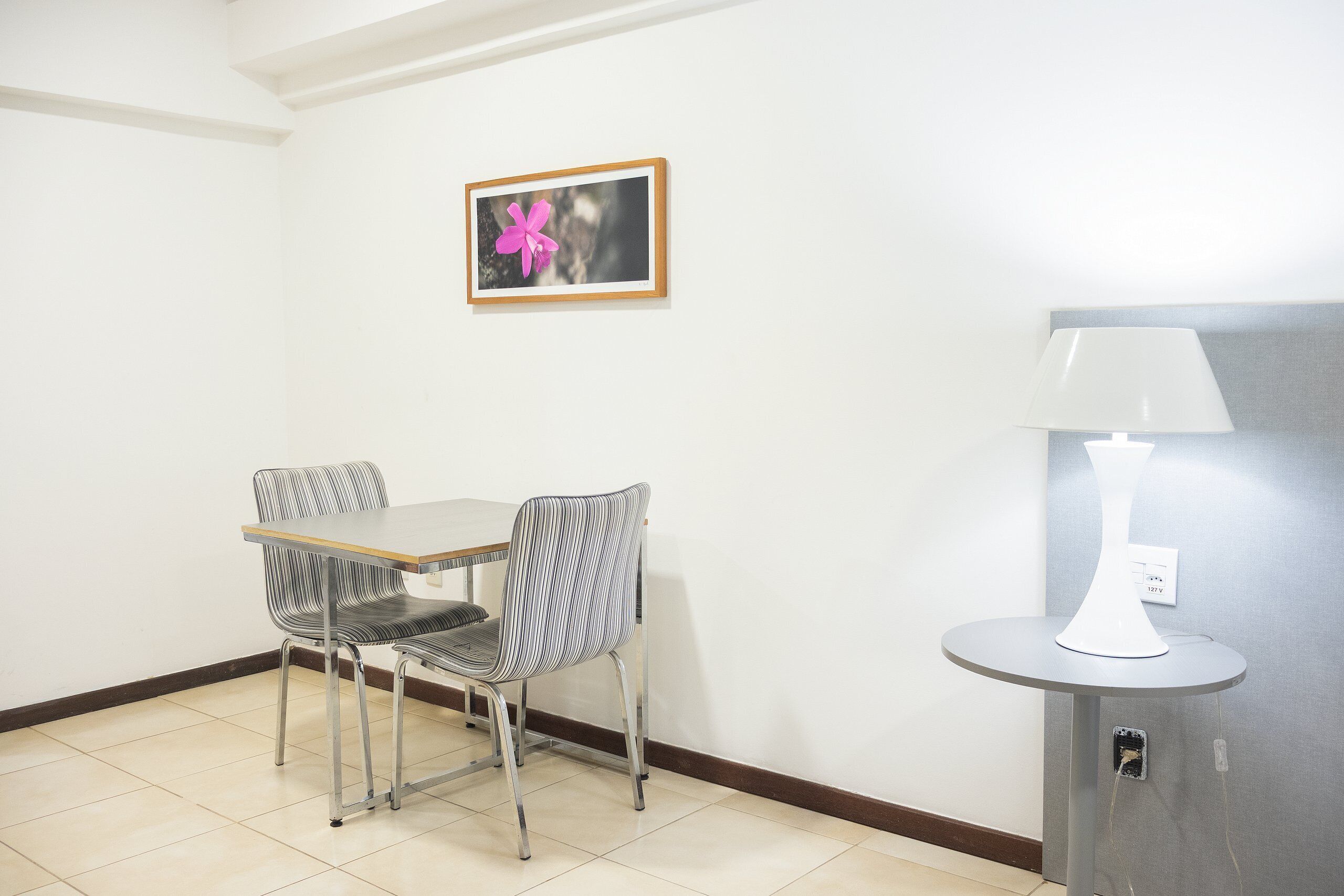 A small dining area with a table and two chairs, a wall-mounted picture, and a lamp on a stand.
