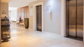 Un moderno vestíbulo de hotel con suelo de baldosas, dos ascensores, un mostrador de recepción y una jaula decorativa.