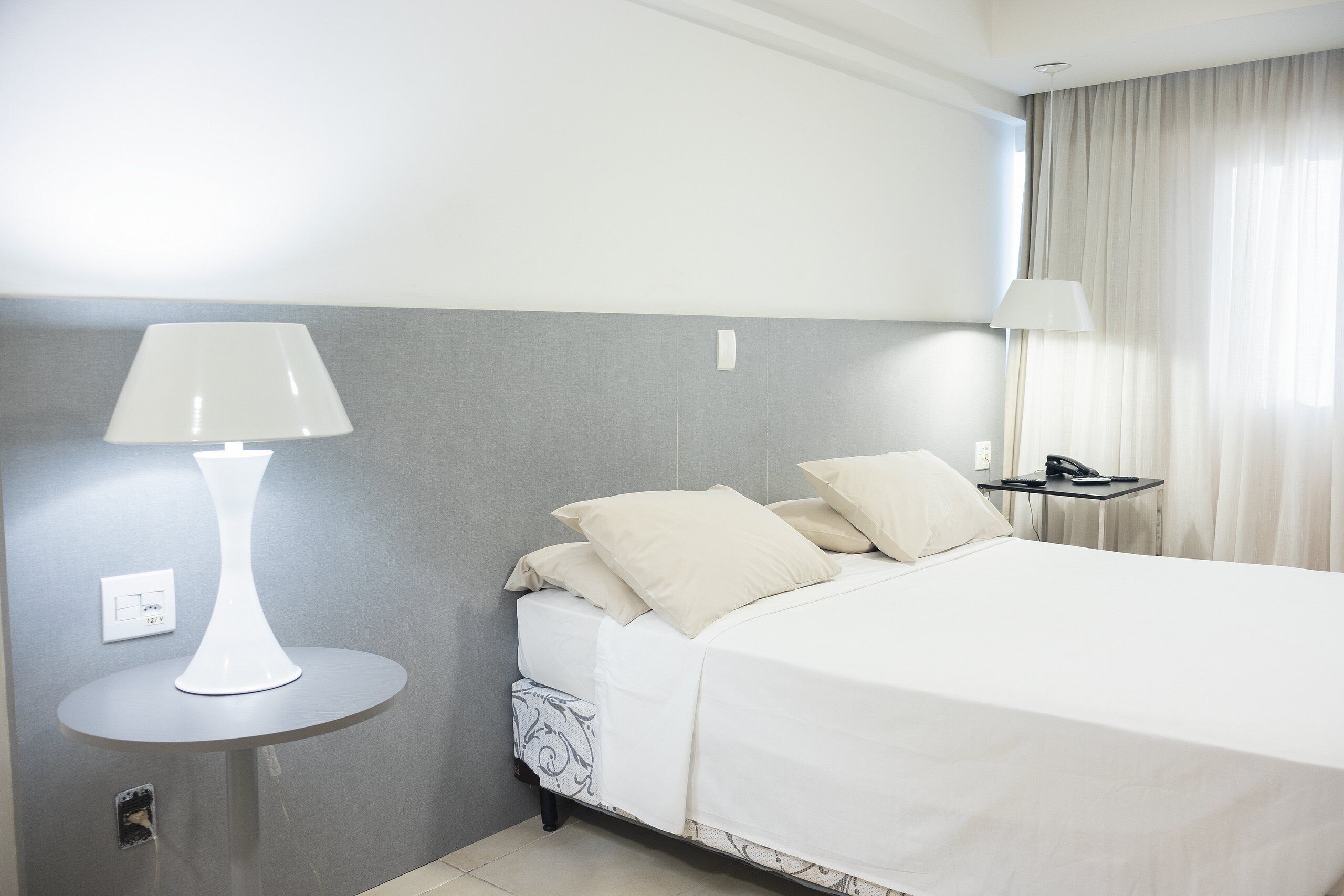 Quarto de hotel com cama, mesa de cabeceira com abajur e criado-mudo com telefone.