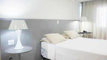 Quarto de hotel com cama, mesa de cabeceira com abajur e criado-mudo com telefone.