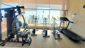 Une salle de sport équipée de divers appareils d’exercice, notamment un tapis de course, un vélo elliptique et des appareils de musculation, et offrant une vue sur la ville grâce à de grandes fenêtres.