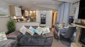 A modern living room with a grey sectional sofa, a dining area with a table set for two, and a kitchen with white cabinets and appliances. - Osprey Court 1 (Great Yarmouth)