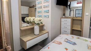 A bedroom with a bed, a bedside table with a vase of flowers, a dresser, a TV mounted on the wall, and a "No smoking or vaping" sign. - Osprey Court 1 (Great Yarmouth)