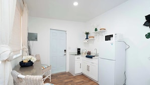 A compact kitchen with white cabinets, a white refrigerator, and a microwave on a white shelf. There is a wicker chair with a striped cushion and a small table with a vase of flowers. - Modern Boho Studio Near Miami River (Miami)