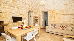Una sala de estar acogedora con una pared de piedra, una mesa de comedor de madera con sillas, un sofá color beige con cojines estampados y un televisor montado en la pared.