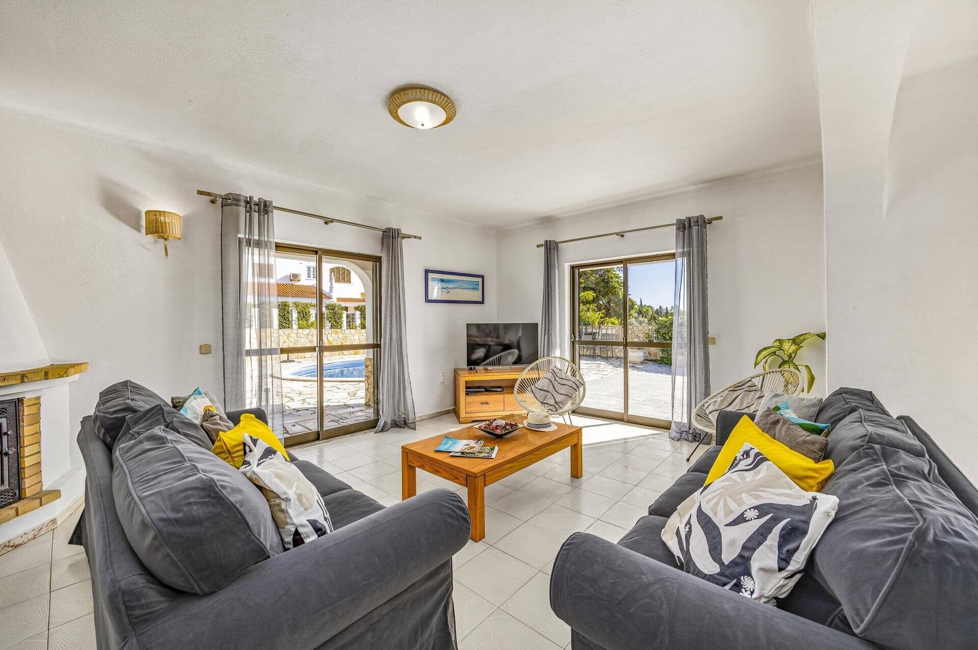 Un salón con un conjunto de sofá, una mesa de centro y un televisor. Hay ventanas grandes con cortinas, una chimenea y vistas a una piscina y vegetación en el exterior.