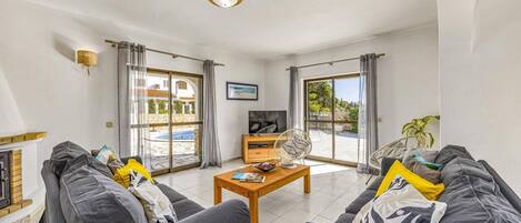 Un salón con un conjunto de sofá, una mesa de centro y un televisor. Hay ventanas grandes con cortinas, una chimenea y vistas a una piscina y vegetación en el exterior.