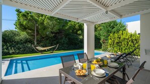 Ein Essbereich am Pool mit einem für das Frühstück gedeckten Tisch, einer Hängematte und Blick auf Pool und Grünfläche.