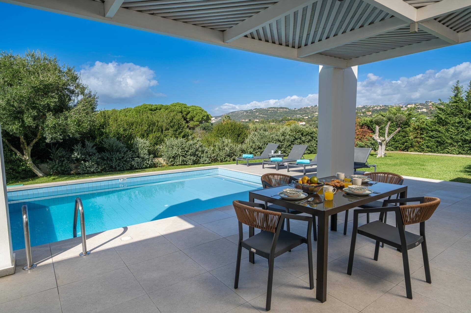 Un área junto a la piscina con una mesa dispuesta para el desayuno, sillones de descanso y vistas del paisaje circundante.