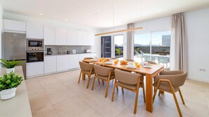 Een moderne keuken met witte kasten, een ingebouwde oven en een magnetron. Er is een eethoek met een houten tafel en zes stoelen.