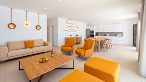 Een moderne woonkamer met een bank, salontafel en oranje stoelen. Er is een eethoek met een tafel en stoelen, en een keuken op de achtergrond.